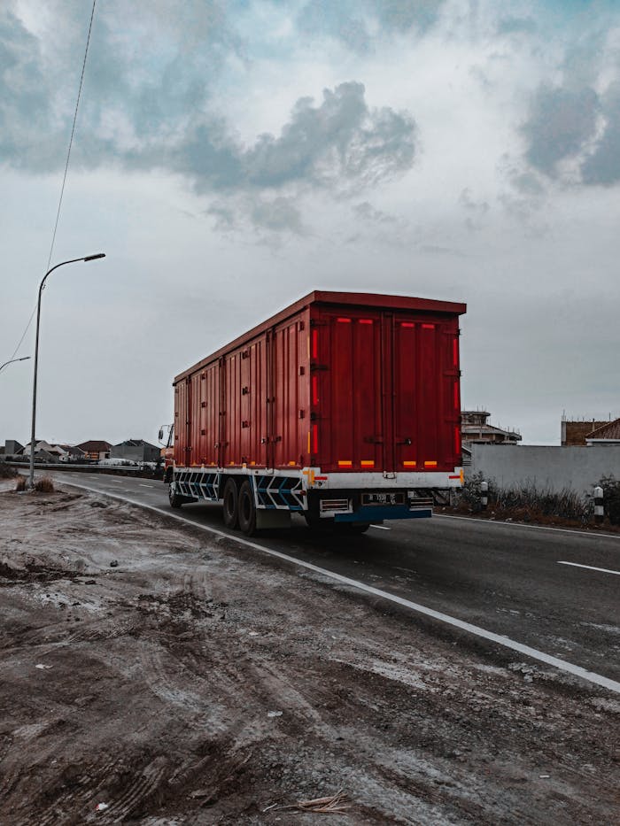 Large heavy motor vehicle driving on roadway while transporting goods along village in cloudy day